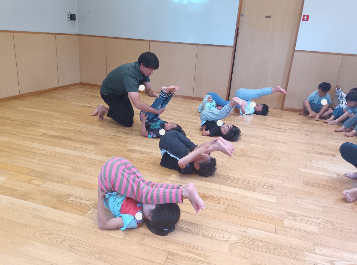 数名の子どもたちが背中を床につけて足を頭の方へあげており、一人の大人は子どもが真っすぐに足が上がるように支えている