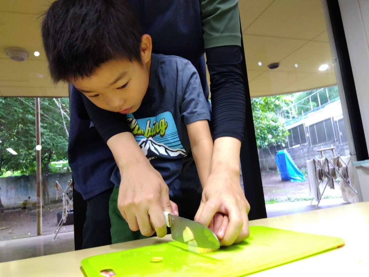 一人の子どもが後ろから大人に手を添えられながらジャガイモを包丁で薄切りにしている
