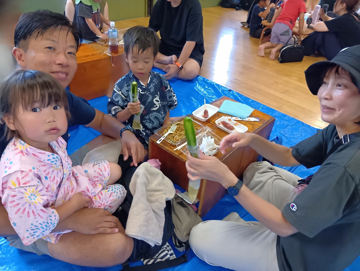 小さい机の上にはフランクフルトと焼きそばが置かれており、二人の子どもと二人の大人がきゅうりの一本漬けを食べいている