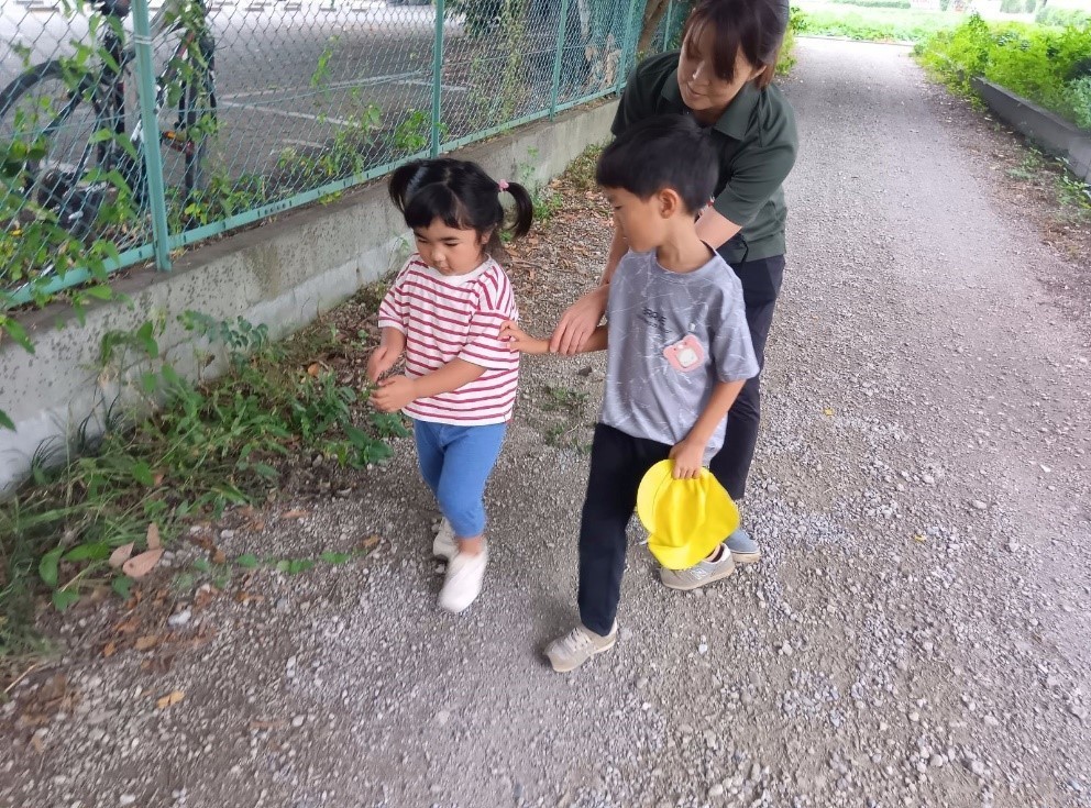 一人の子どもが隣の子どもの肩に手を伸ばして触れており、大人が後ろからその手を支えている