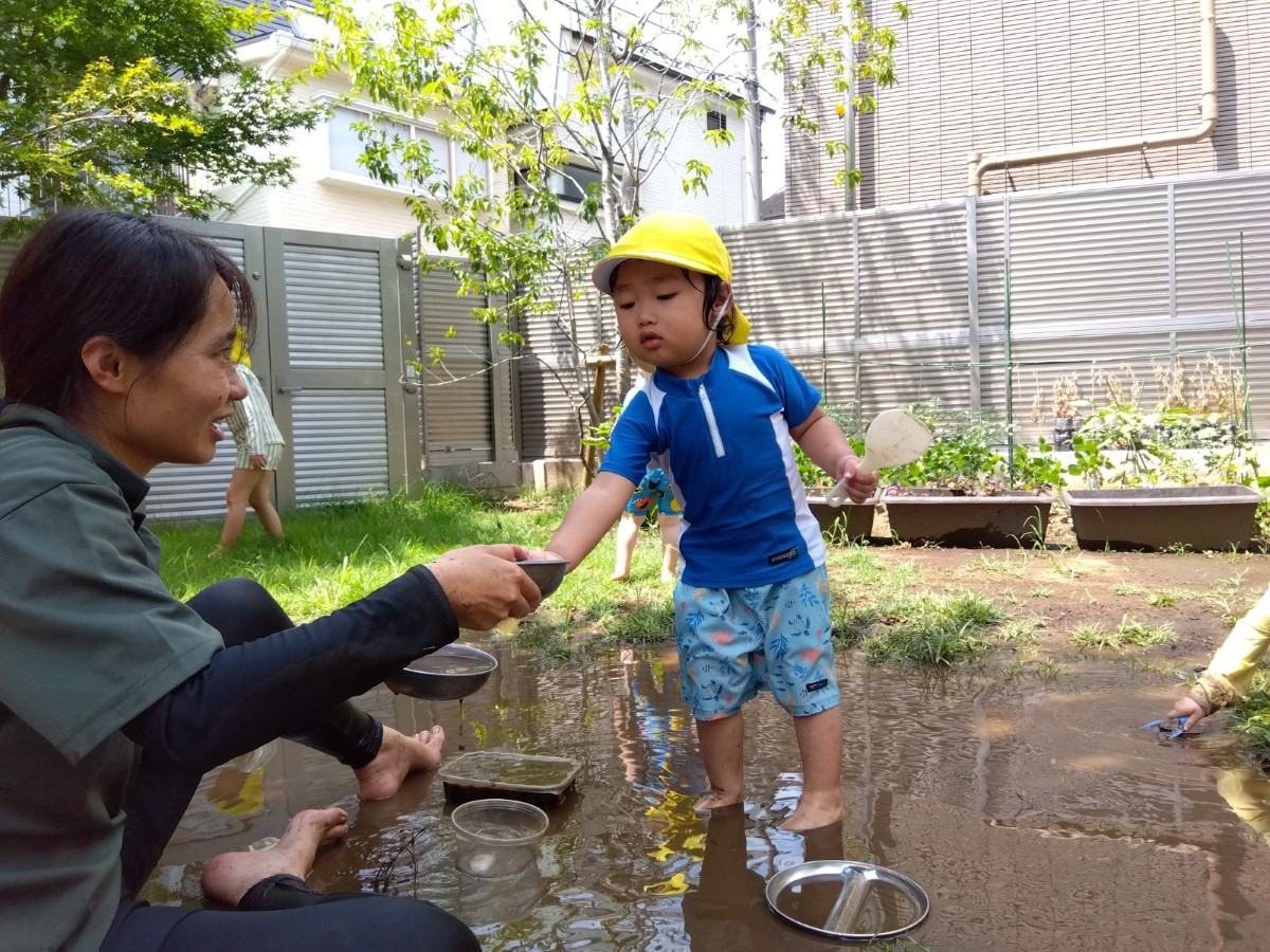 泥遊びで、裸足で泥の中に立つ子どもが片手にしゃもじを持ち、大人から泥の入ったお椀をもらおうとしている