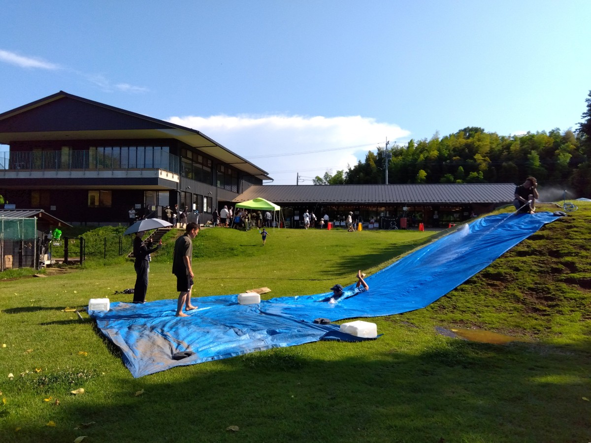 築山にブルーシートが敷かれたスライダーでは、上から大人が水を流し、一人の子どもが滑り降りている