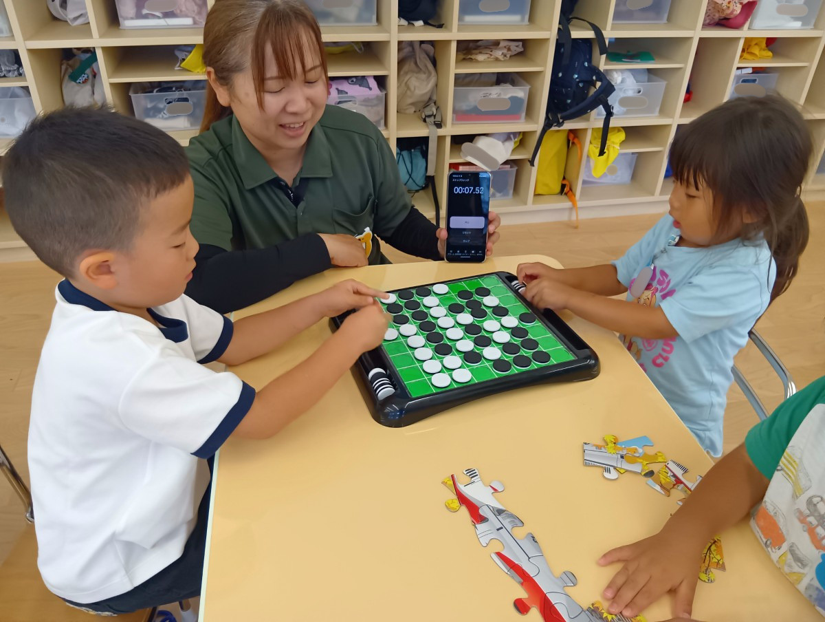 机上では、二人の子どもが向かい合ってオセロで遊んでおり、大人が傍で時間を計っている。同じ机の隣の子どもはパズルで遊んでいる。