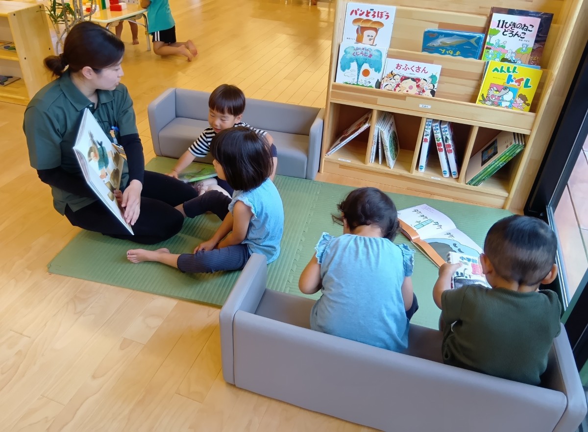 絵本コーナーにはマットが敷かれ長椅子が置かれており、二人の子どもが椅子で絵本を読んでいる。マットの上では一人の大人が二人の子どもに絵本の読み聞かせをしている。
