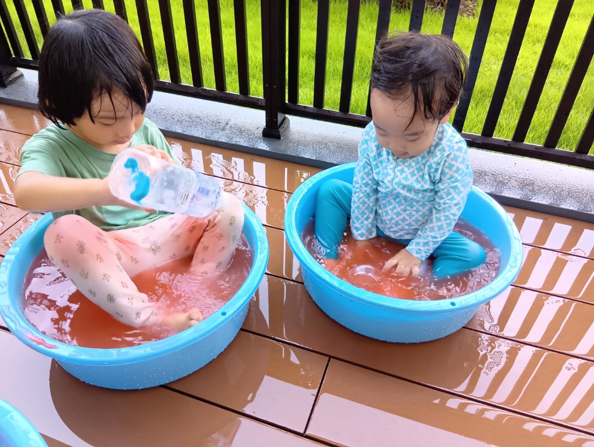 二人の子どもが色水が入ったたらいの中にそれぞれ入り、色水が入ったペットボトルを流してたらいの色水を見ている