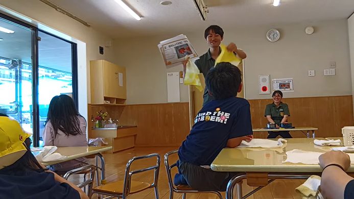 一人の大人が色付きの布と画用紙を手に持って前に立っており、子どもたちは白布が置かれた机の前に座話をって聞いている
