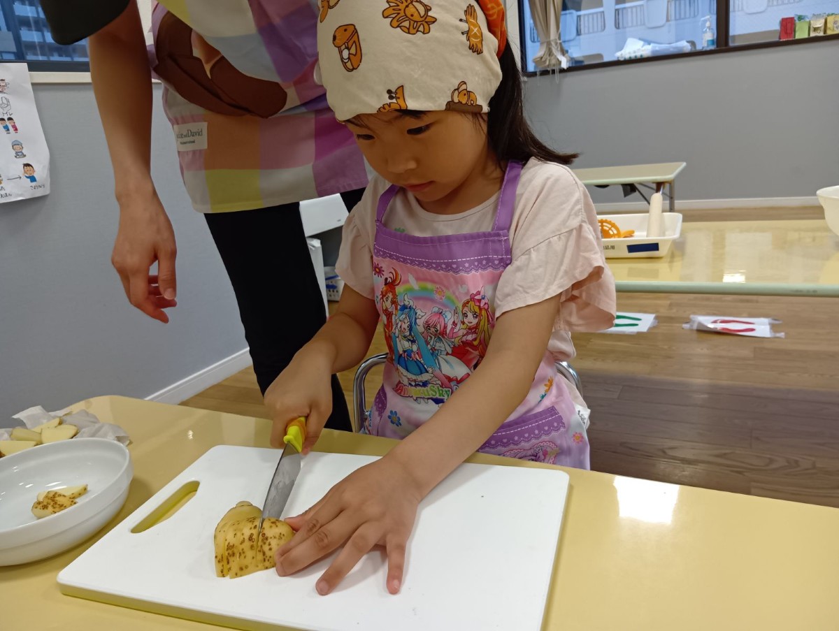 エプロンと三角巾を身に着けた子どもが包丁を使ってジャガイモを切っている