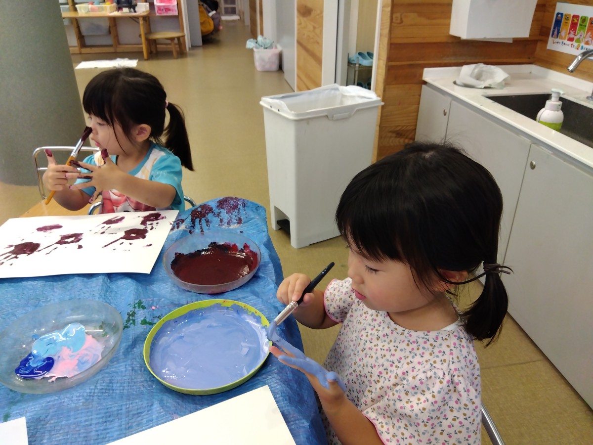 椅子に座った二人の子どもが、筆を使って自分の掌に絵の具を塗って画用紙に手形を付けている