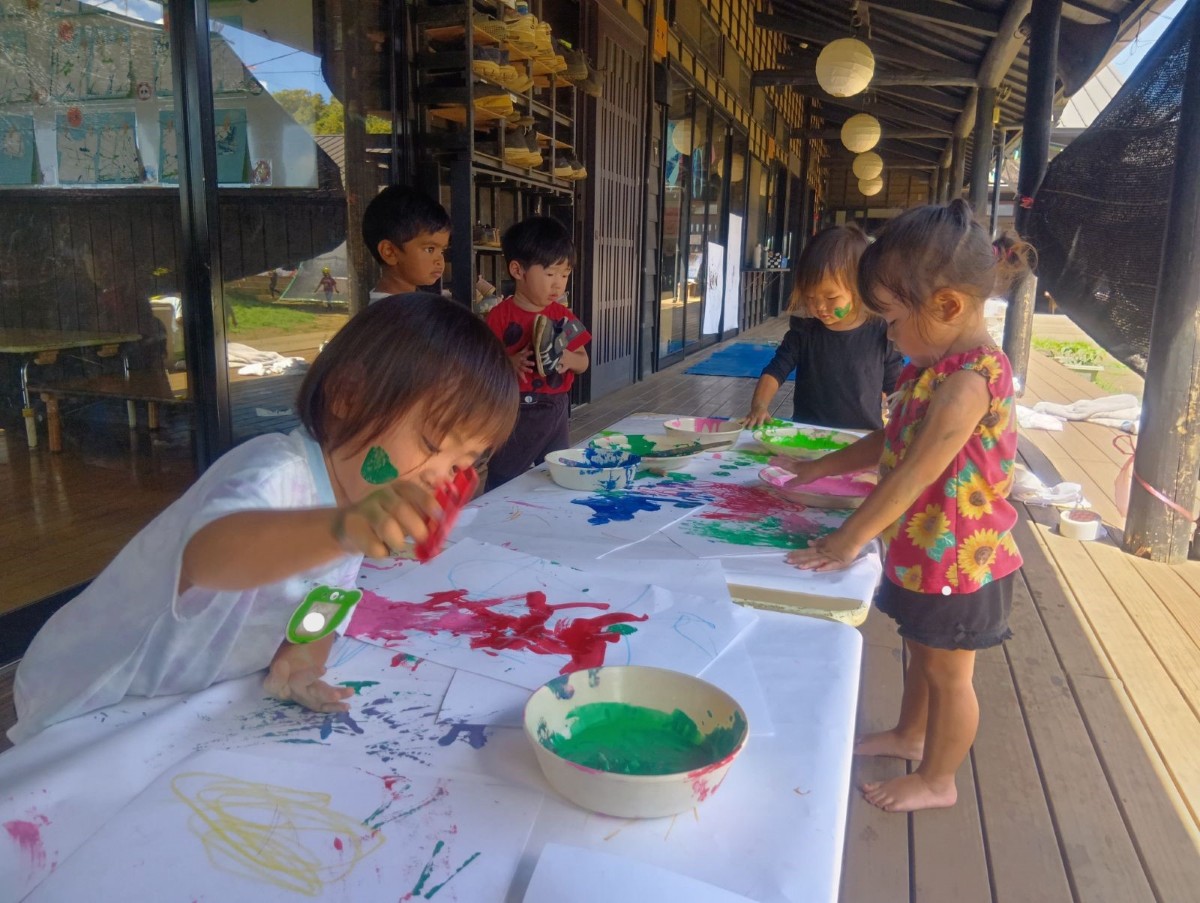 縁側にある机の上に白画用紙と絵の具が入った皿が置いてあり、子どもたちは顔に絵の具を付けたり洗濯バサミでスタンプしたり手形をつけたりしている