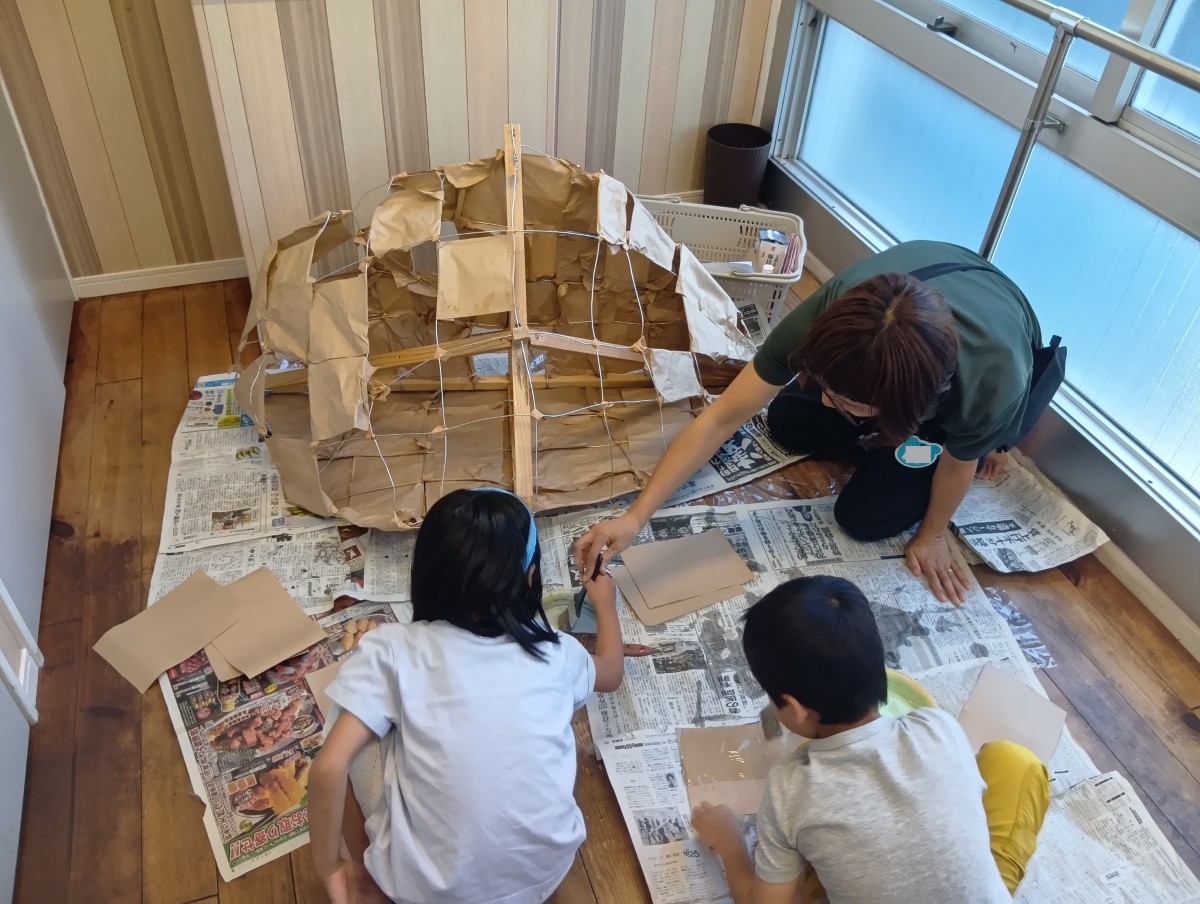 新聞紙が敷かれた床には、中が空洞の木とワイヤーの骨組みに紙が途中まで貼り付けてあり、子どもが刷毛を使って紙に糊を着けている