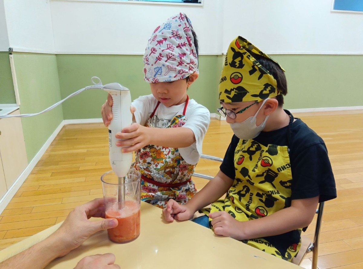 一人の子どもがブレンダーを持ちトマトを混ぜ、隣の子どもはその様子を見ている