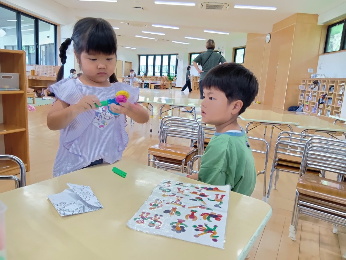もう一人の子どもが机の傍に立ち、途中までくっついたブロックを持って続きを作ろうとしており、先程と同じ子どもがその様子を見ている