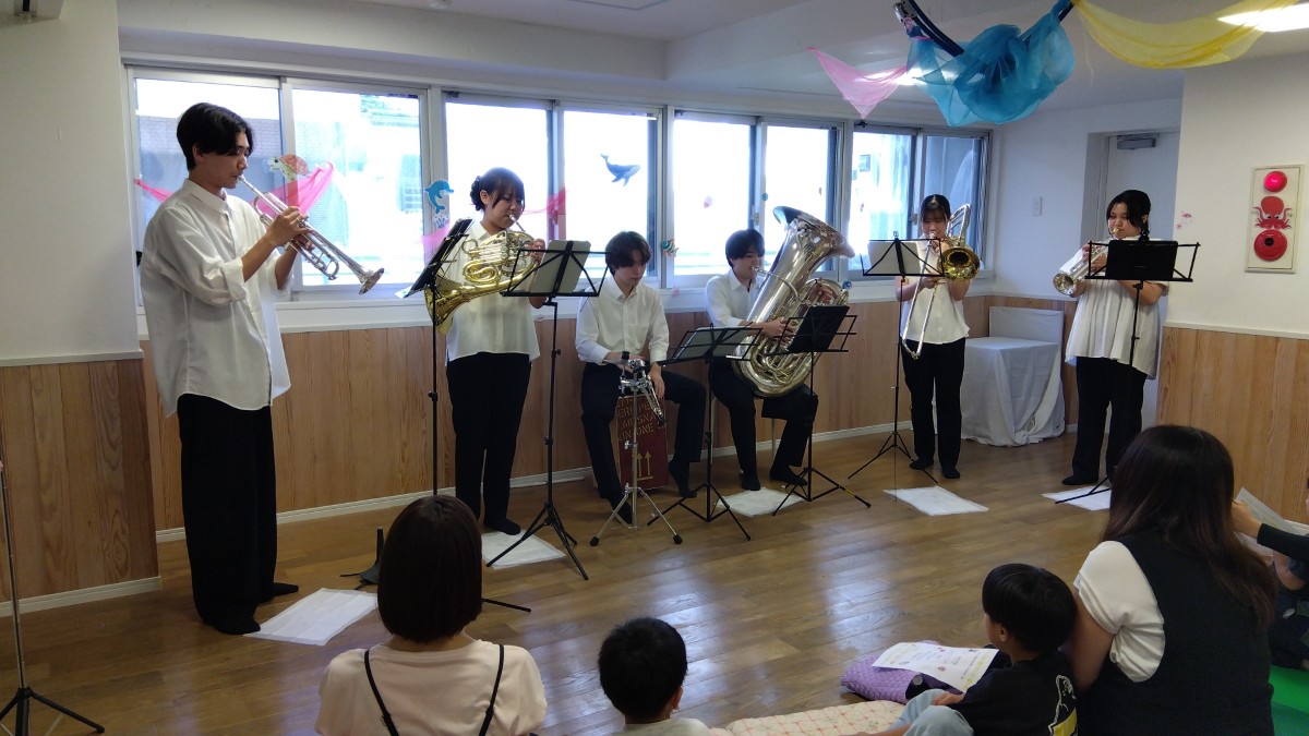 前で学生たちが管弦楽を演奏しており、それを大人と子どもたちが座って聞いている