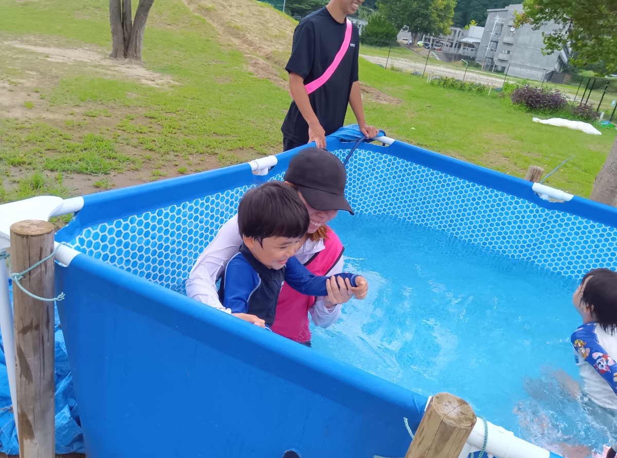 プールでは、一人の大人が一人の子どもを抱き上げて水から出しており、周囲の子どもたちはその様子を笑顔で見ている