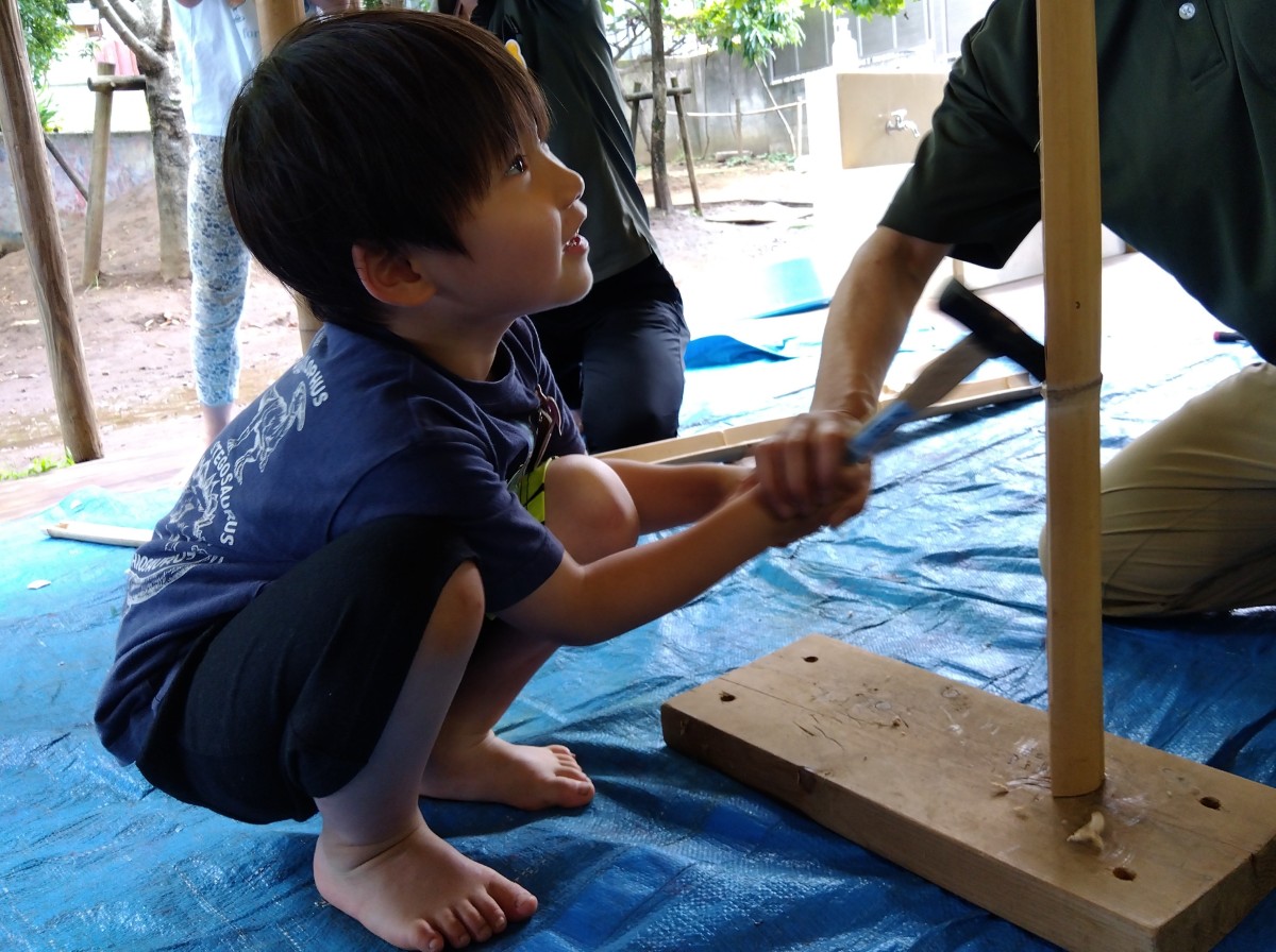 一人の子どもが大人に手を添えられながらトンカチを握り、しゃがんで竹を叩いている
