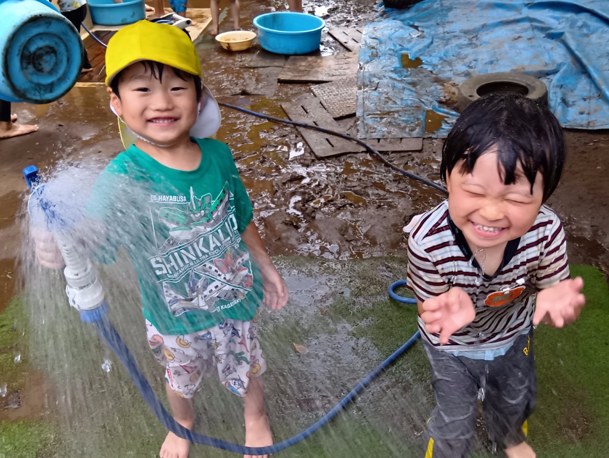 一人の子どもが水が出ているホースを手に持ち、隣の子どもは水でびしょぬれになって笑っている