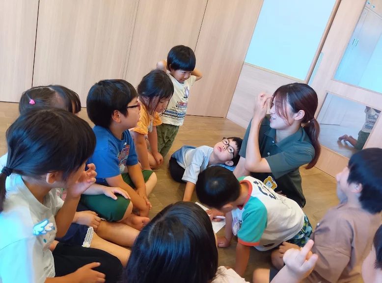 一人の大人がペンと紙を持ち、その周囲に座っている子どもたちと話をしている