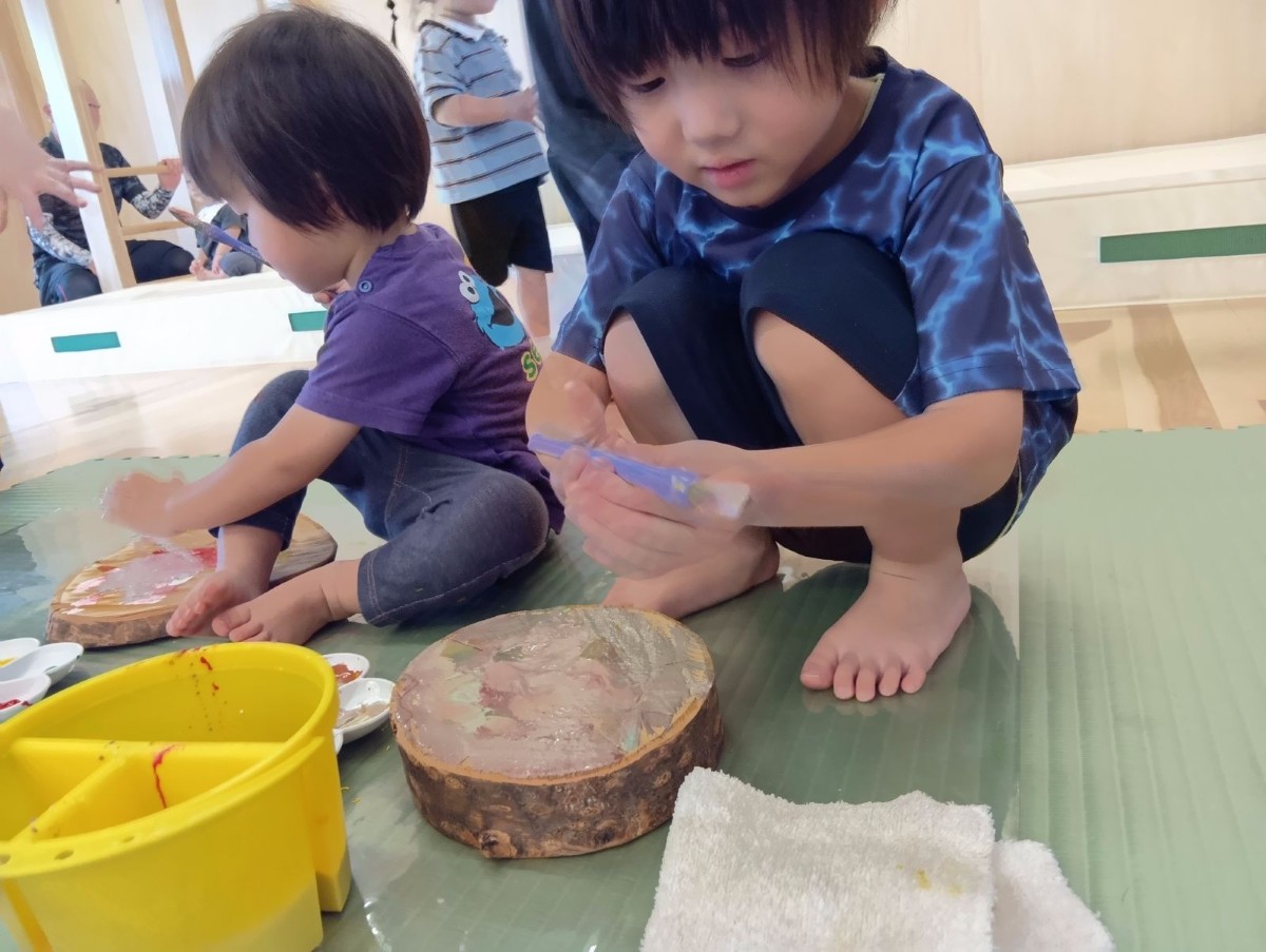 室内では、絵の具、筆洗い、切り株の前に子どもが筆を持ってしゃがんでいる