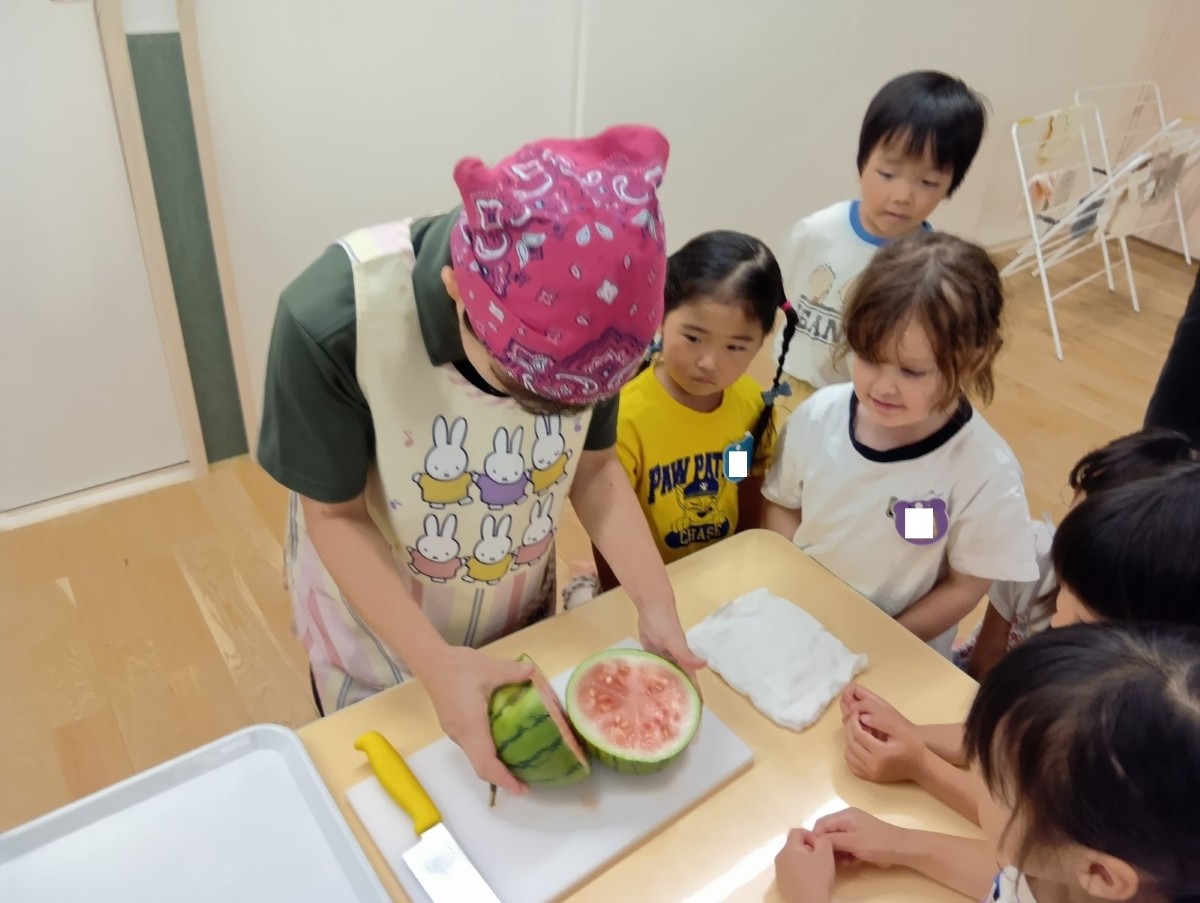 エプロン三角巾を着けた大人が、大人の掌サイズのスイカを包丁で半分に切っており、その様子を子どもたちが周りで見ている