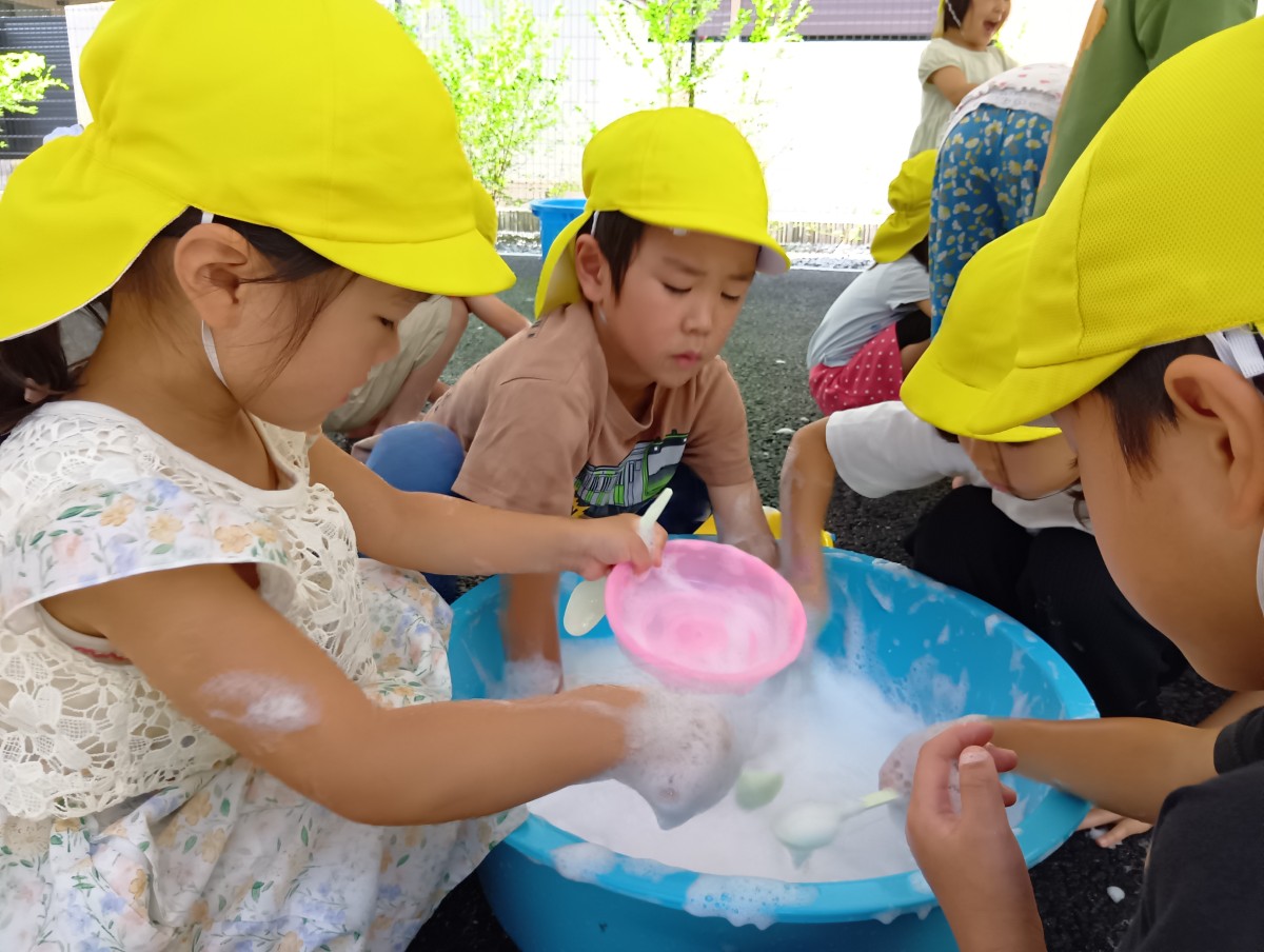 子どもたちは、泡がたくさん入ったたらいの中を囲み、カップやスプーンで泡を掬ったり直接手で触れたりしている