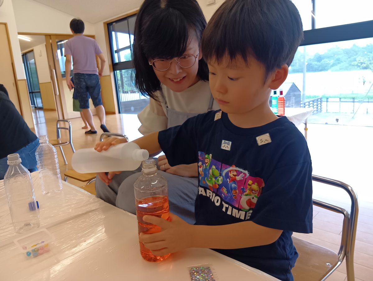 一人の子どもが色水が入ったペットボトルに液体を丁寧に入れており、すぐ傍に大人がその様子を笑顔で見守っている
