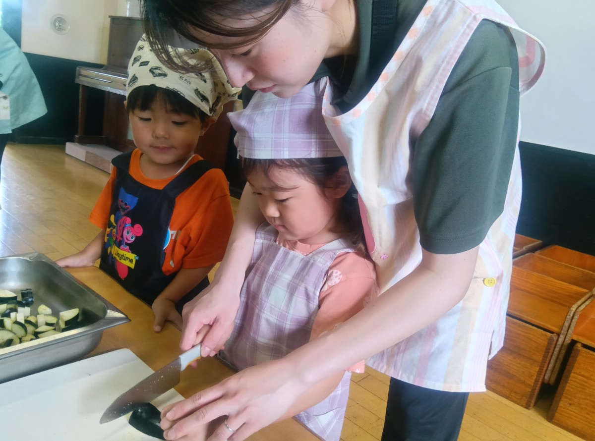 エプロン三角巾を着けた子どもが後ろから大人に手を添えられながらナスを包丁で切っている