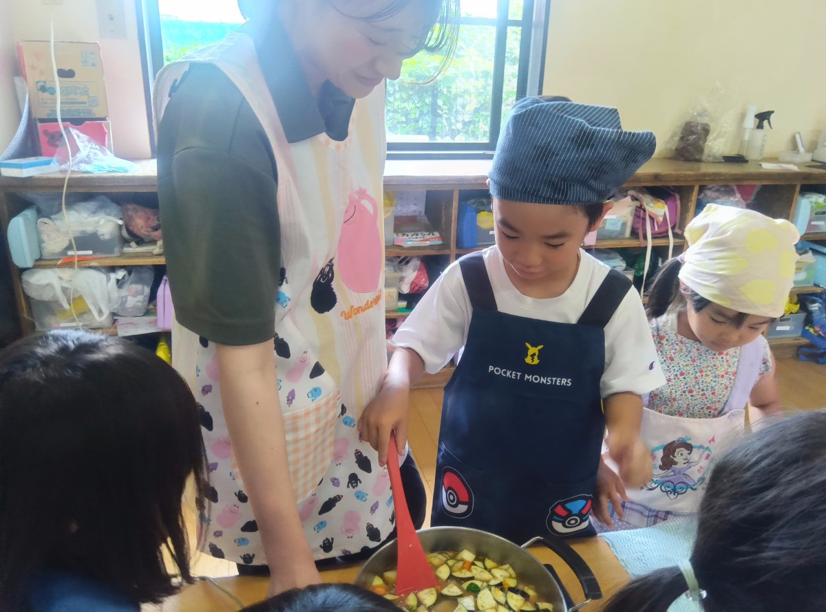 エプロン三角巾を着けた子どもが鍋の中に入っている切られた野菜をヘラで混ぜている
