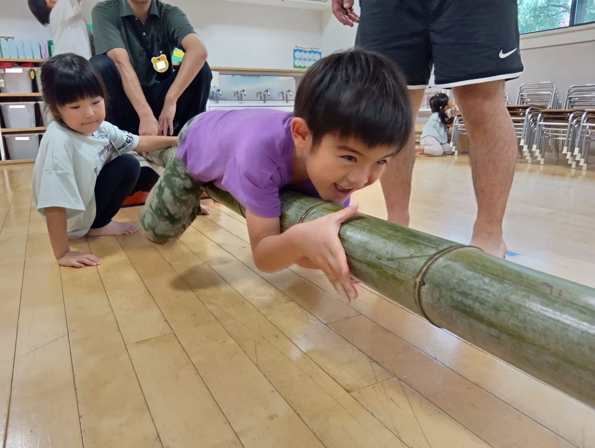 室内では、一人の子どもが平均台に見立てた竹に寝転んで抱き着き、手足を使って前に進もうとしている