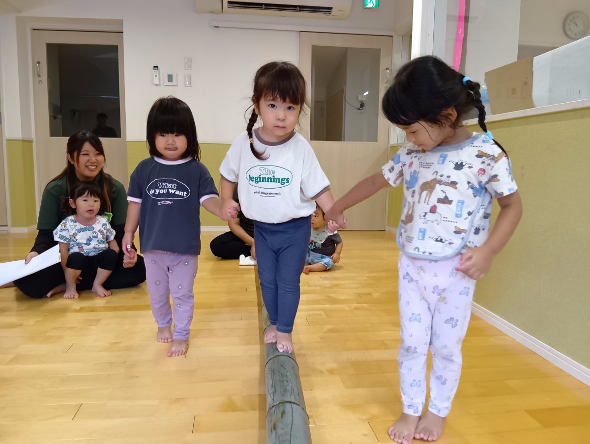 室内には平均台に見立てた横になった竹があり、竹の上を歩く子どもとその子どもの右手、左手と手をつなぐ二人の子どもがいる