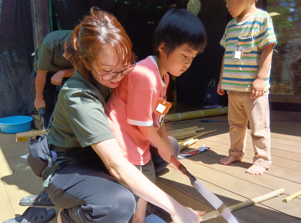 一人の子どもが後ろから大人に身体と竹を支えられながら、細い竹をのこぎりで切っている