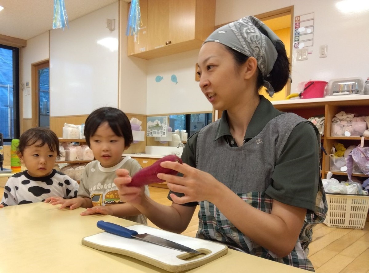 包丁とまな板が置かれている机の前で、エプロン三角巾を着けた大人が、さつまいもを手に持って子どもたちに見せている