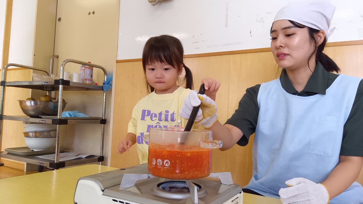 コンロの上にはつぶれたトマト液が入った鍋をコンロの火にかけ、大人と子どもがお玉を一緒に持ち、液体を混ぜている