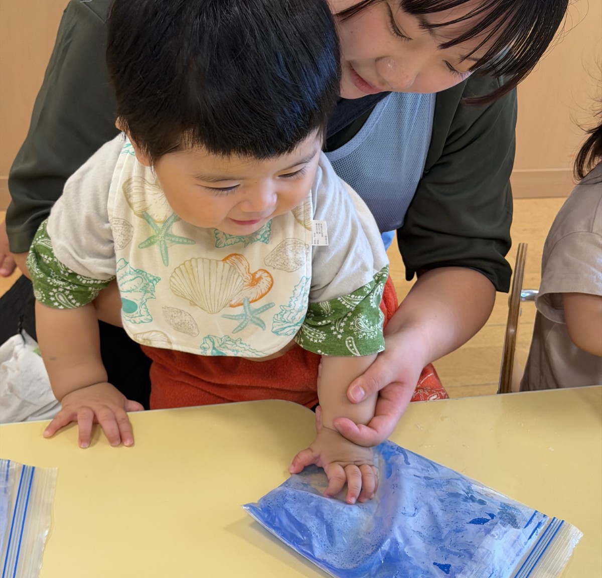 一人の子どもが大人に手を添えられながら、青い液体の入ったジッパー付きビニール袋に掌で触れている