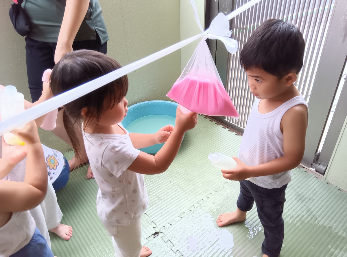 一人の子どもが紐にぶら下がっている色水が入ったビニール袋の下に容器を近づけており、もう一人の子どもは容器を持って順番を待っている