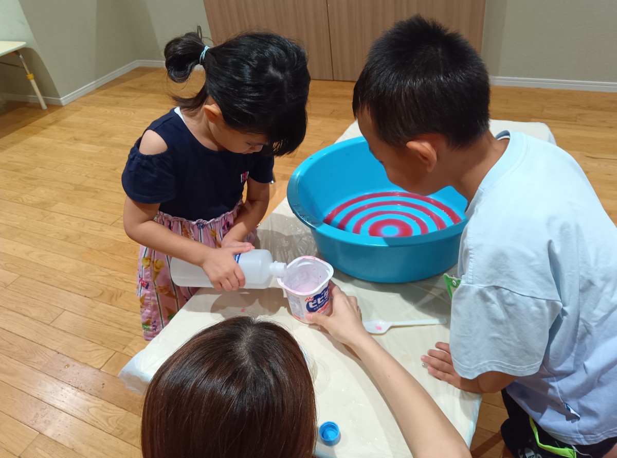 一人の子どもが液体を器に入れようとしており、そばでもう一人の子どもがその様子を見ている