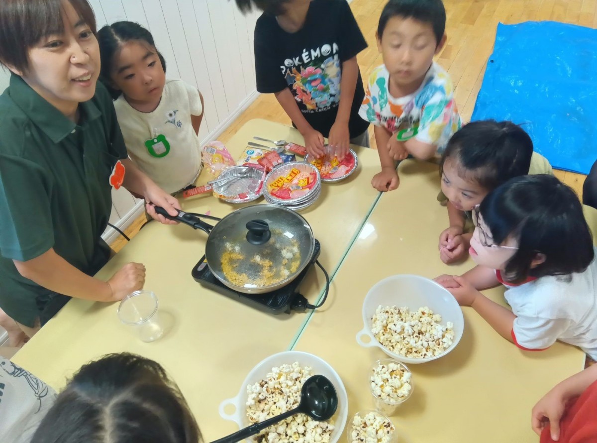 机の上で大人がフライパンを持ってポップコーンを作っており、その周りに子どもたちが集まって様子を見ている