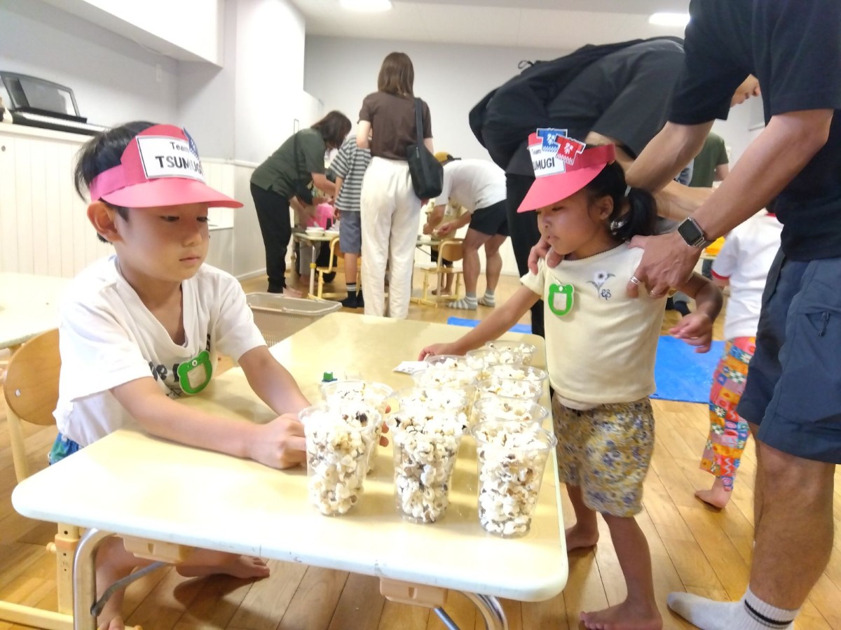 ポップコーンが入った透明カップがたくさん並んでいる机の前に、サンバイザーを着けた子どもが机を挟んで両側にいる