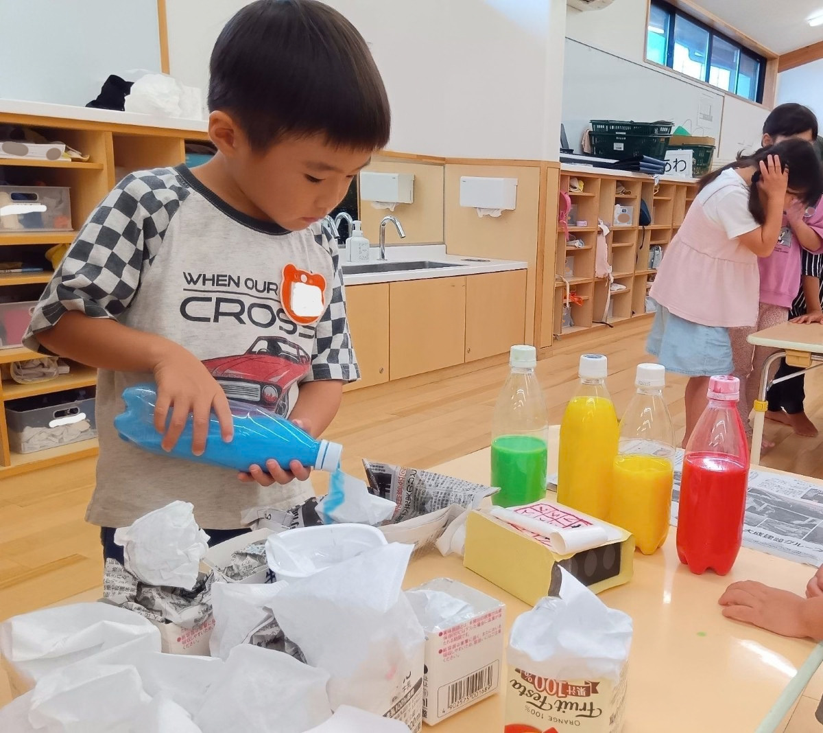 色水が入ったペットボトルが並んだ机では、一人の子どもが水色の色水が入ったペットボトルを手に取り、廃材で作られたかき氷に液体をかけている