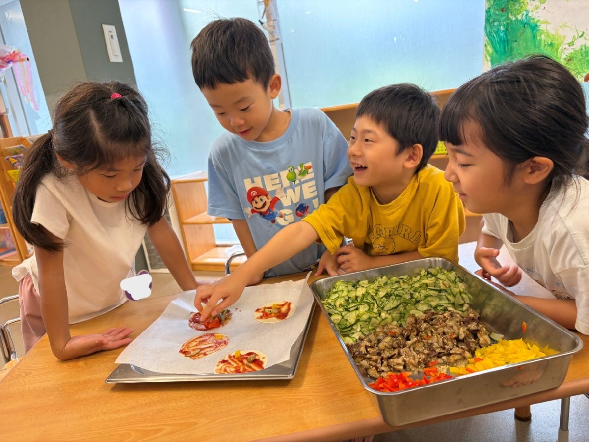 ケチャップが塗られた生地の周りに子どもたちは集まり、その中の一人の子どもが具の載っているピザを指差している