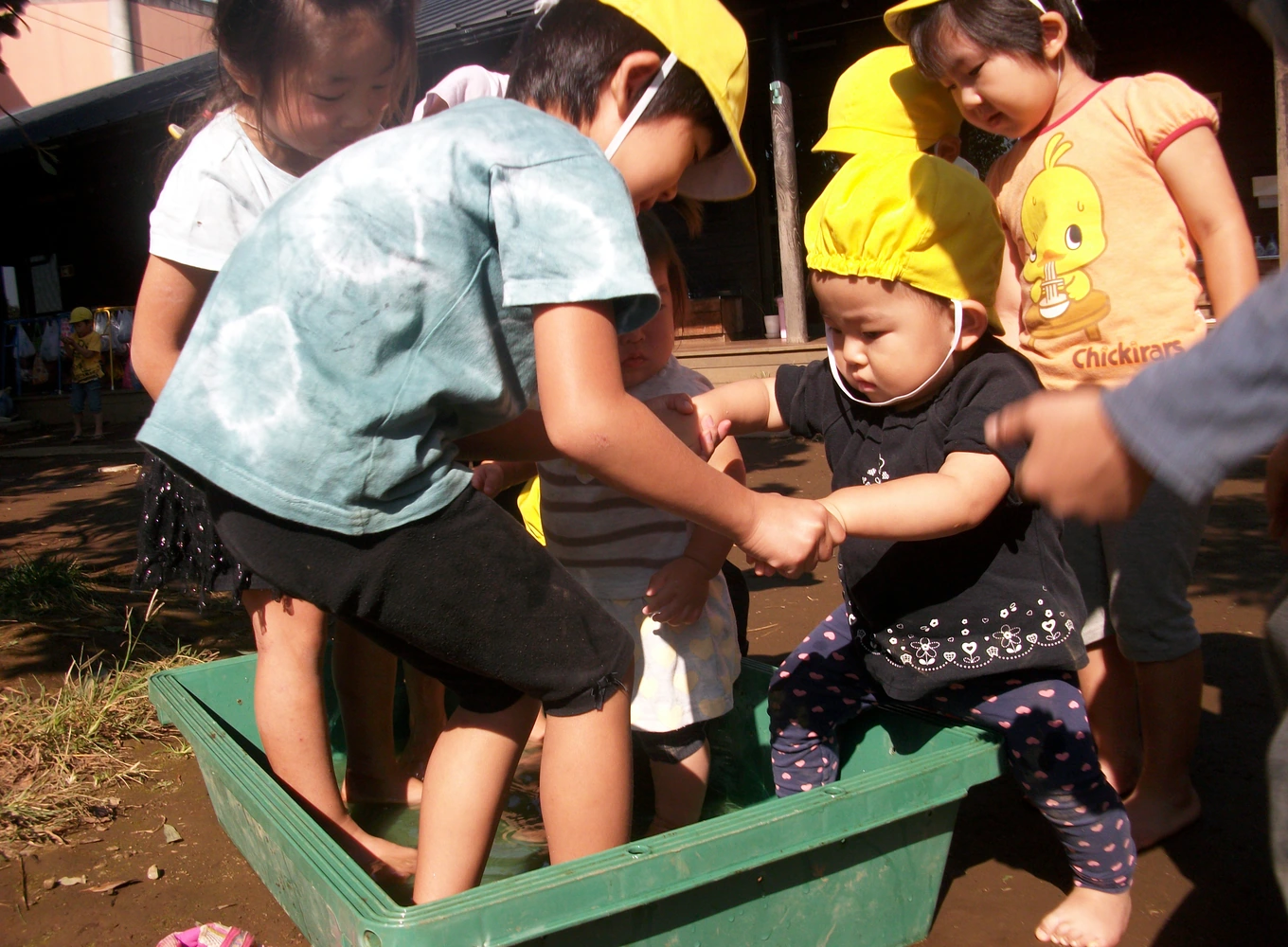 An older child helping a younger child step into a sandbox