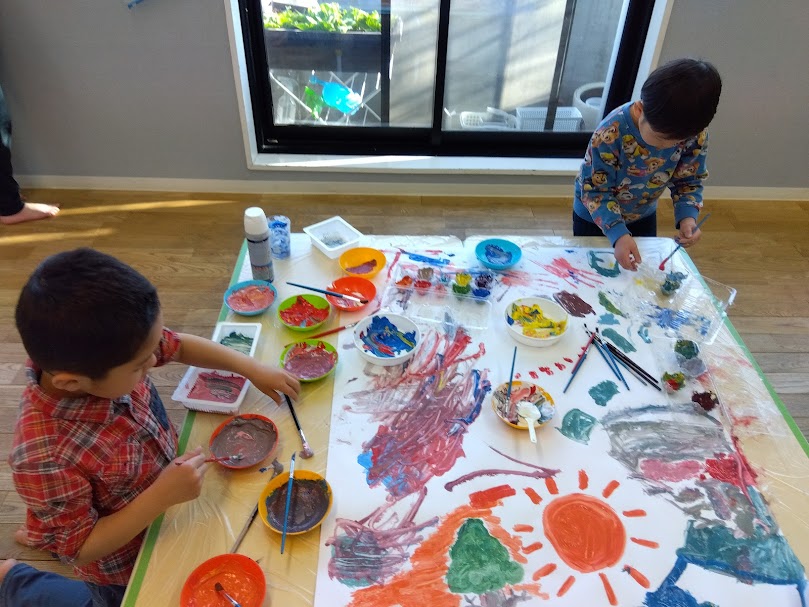 机の上に色とりどりの絵具を用意して紙に絵を描く子どもたち