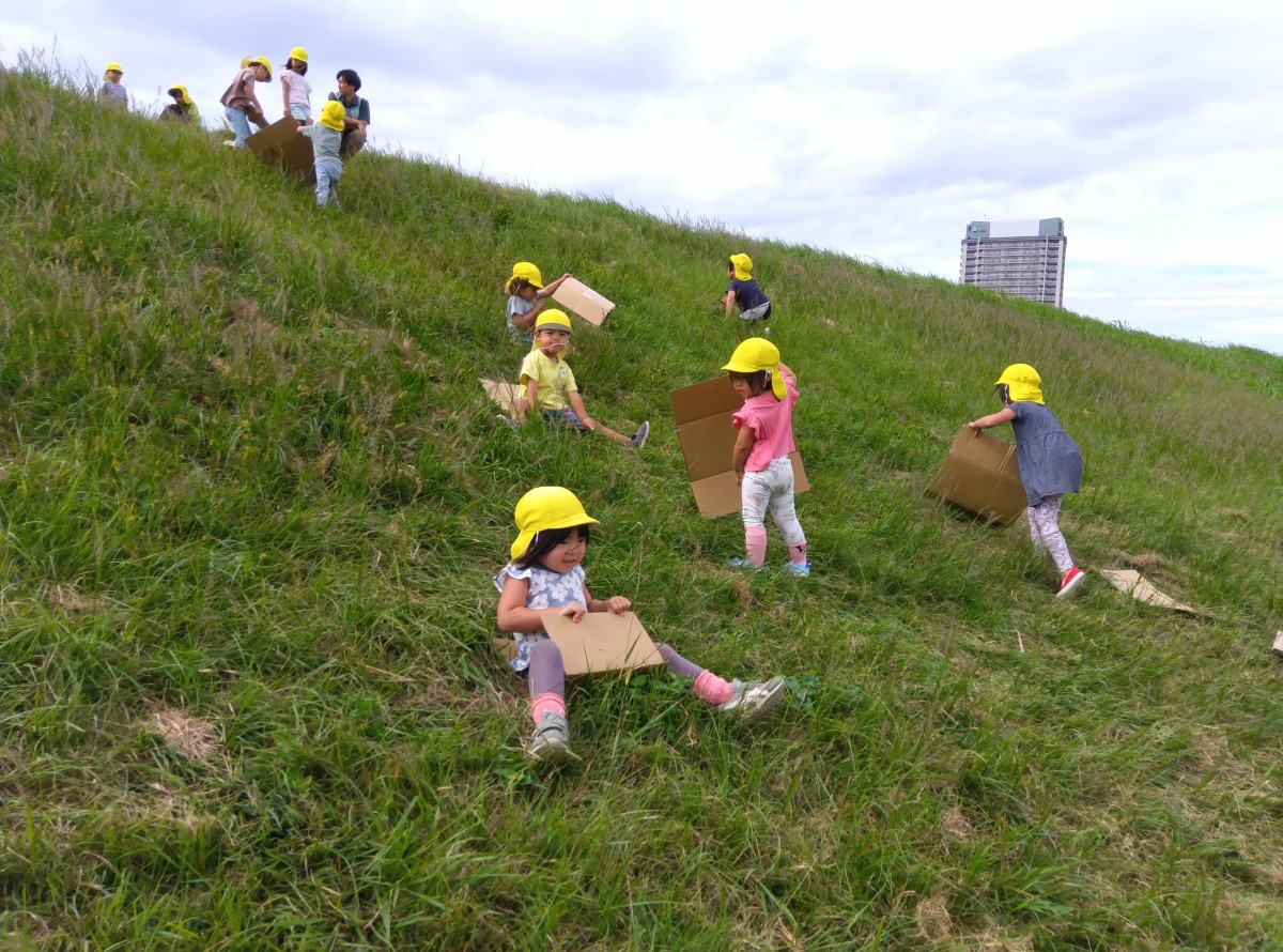 土手で段ボールを使った芝滑りを楽しむ子どもたち