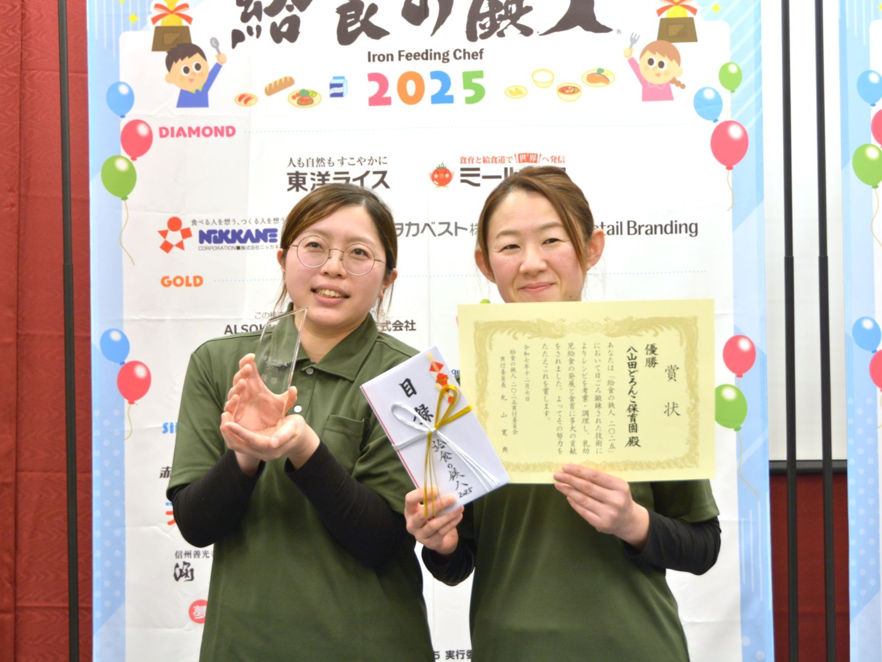 給食の鉄人参加者の二人が優勝トロフィーと賞状を持って記念撮影に臨む様子