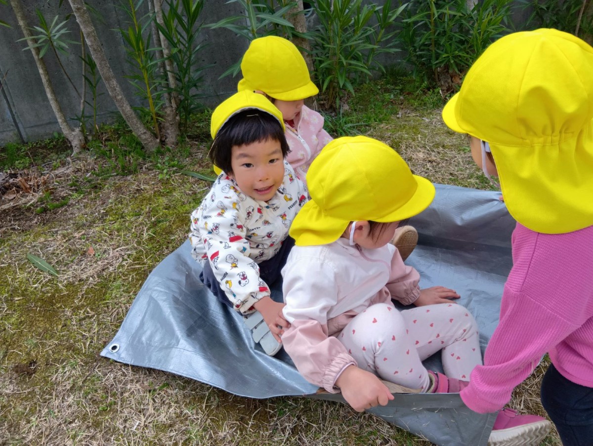 レジャーシートの上に３人の子どもが座り、一人の子どもがシートを引っ張り芝滑りをしている