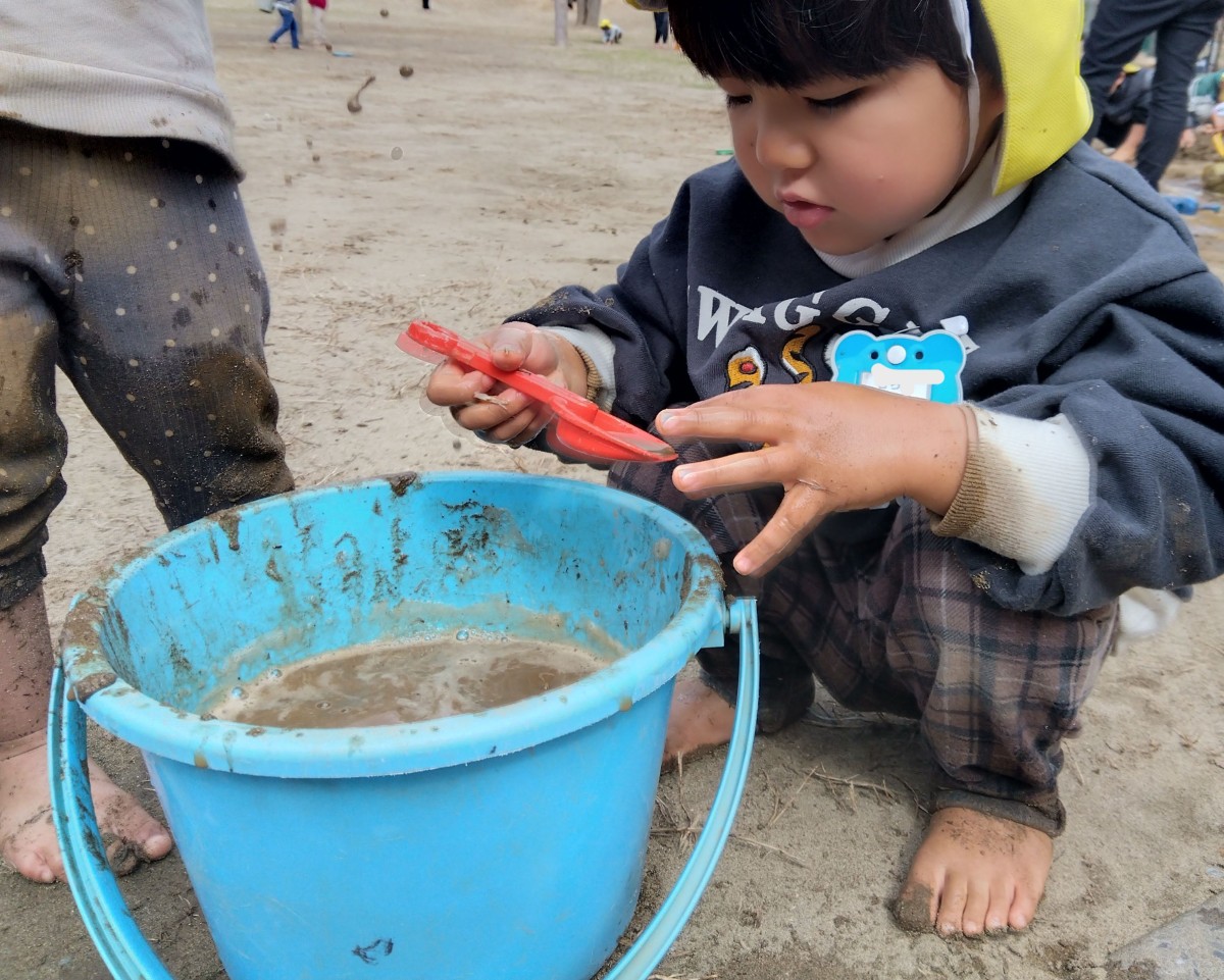 子どもがバケツに入った泥水をスコップですくい、指先で泥水に触れようとしている
