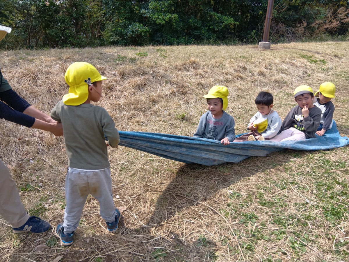 一人の子どもが大人と一緒にブルーシートの端を持ち、友達が座って乗っているそのブルーシートを引っ張っている