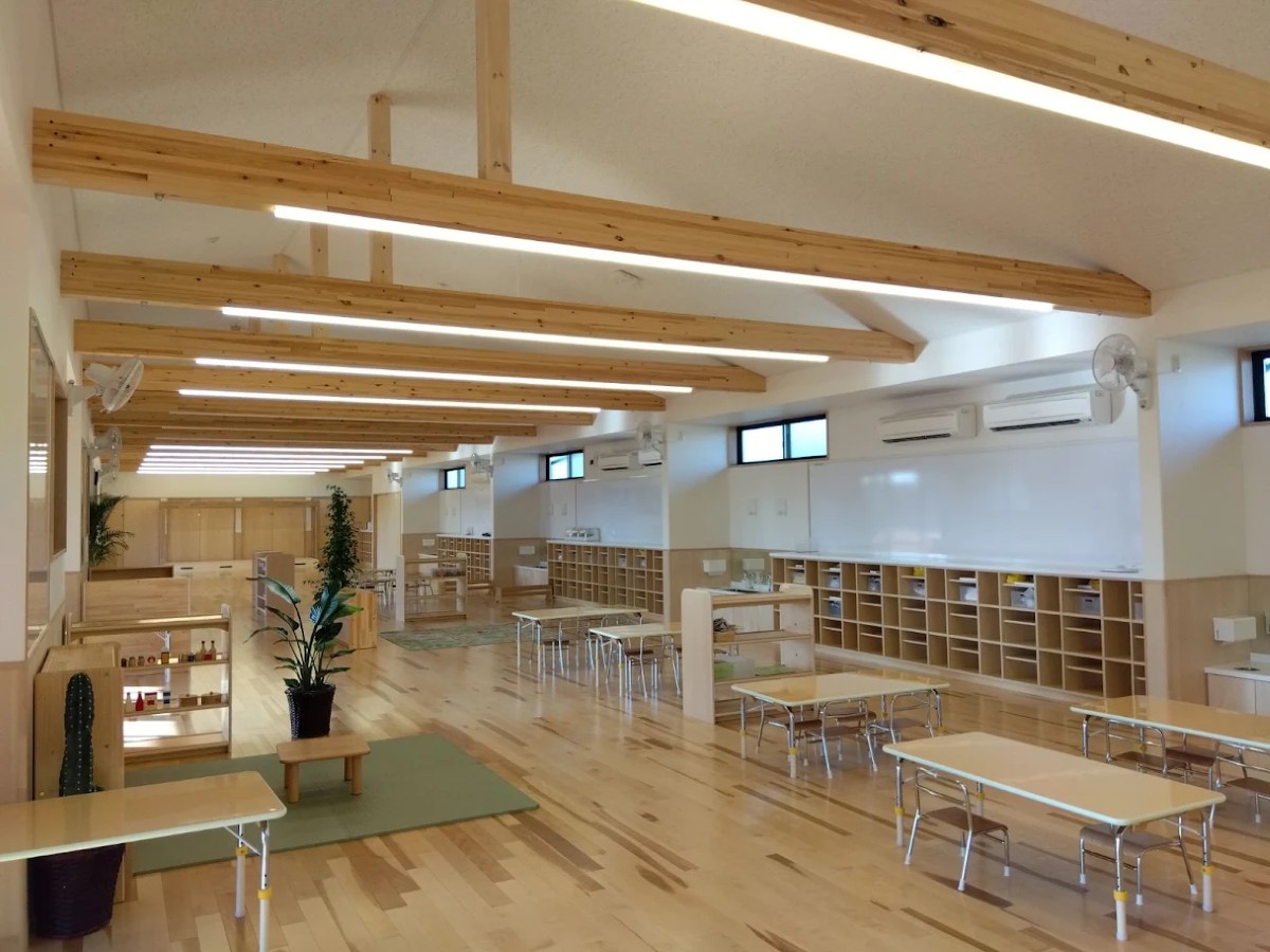 保育室には幼児用の机と椅子が並び、壁側にロッカー、幼児用玩具が入った棚、植物などが置かれている
