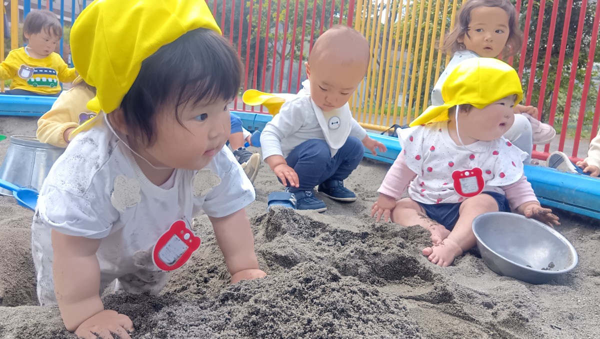 公園の砂場にはバケツやシャベルが置いてあり、はだしで砂場に入り砂に触れる子、靴を履いて砂場にしゃがみ込む子、砂場の外で様子を見ている子などがいる
