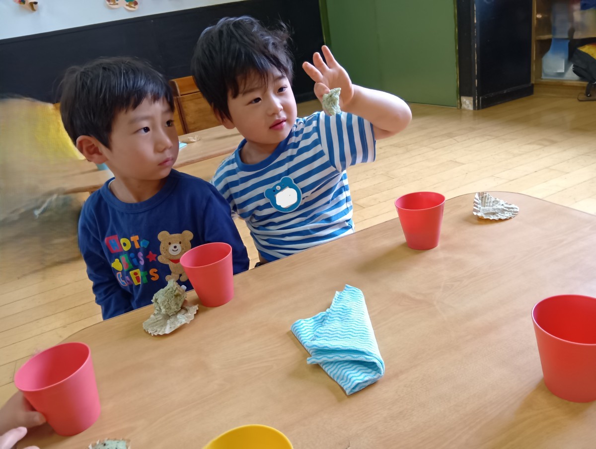 一人の子どもがおやつのよもぎ蒸しパンを指でつまみあげ、隣の子どもと一緒に蒸しパンを見つめている。