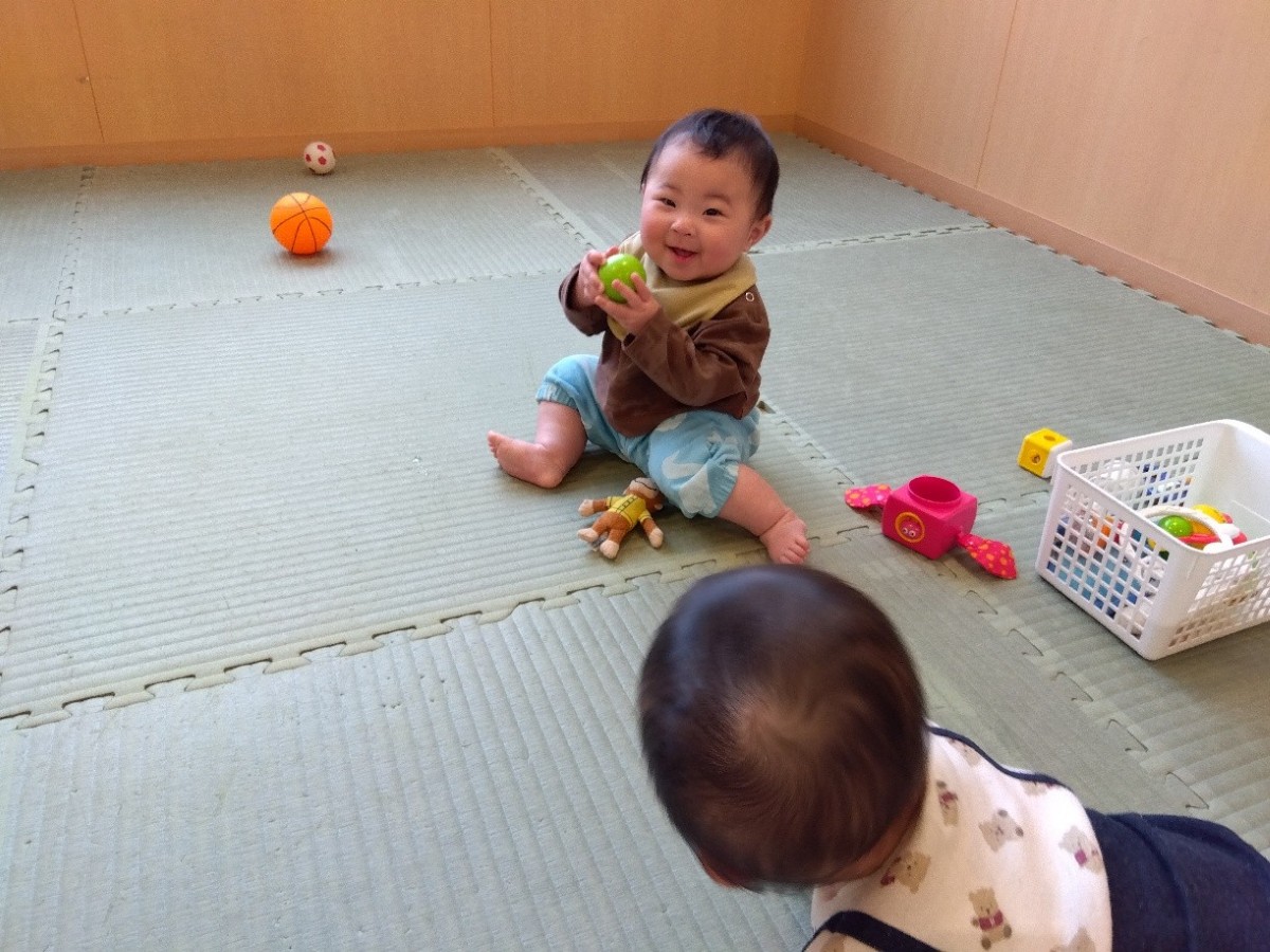 室内で玩具遊びをしている乳児が、両手にボールを持ち、笑顔を見せている。